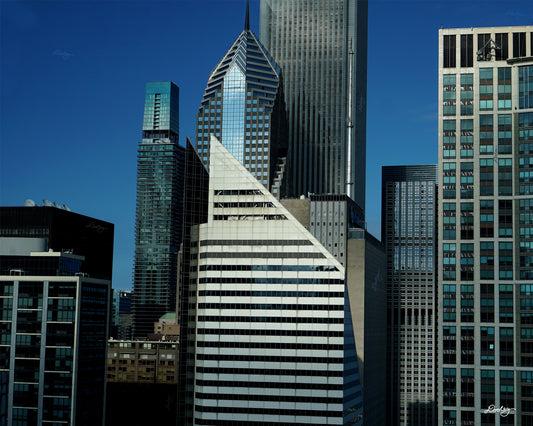 Image showcasing a cluster of towering skyscrapers in Chicago, with a vibrant blue sky as a backdrop. The buildings exhibit a variety of architectural styles, with sleek glass facades and modern designs dominating the skyline. A particularly striking feature is a diamond-shaped skyscraper, Crain Communications Building, with its unique silhouette standing out amidst the other buildings. Buildings also include Two Prudential Plaza, The St. Regis Chicago, and a host of smaller buildings.